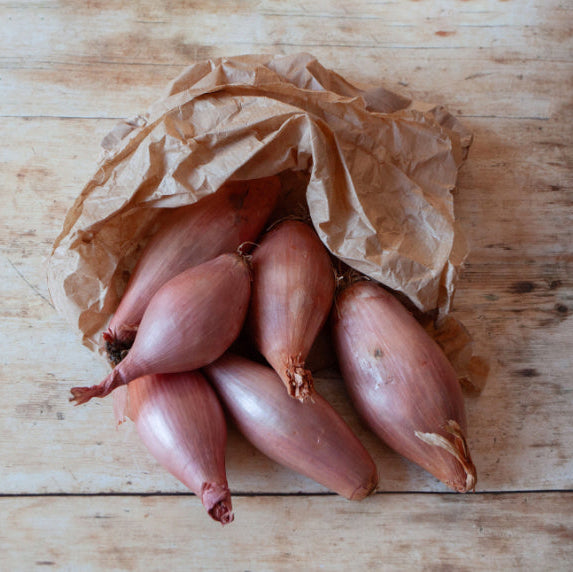 A group of shallots spilling out of a brown paper bag onto a wooden surface.