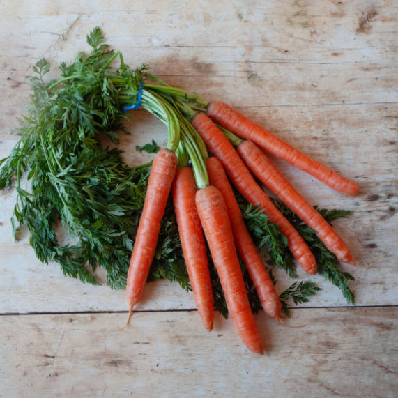 A bunch of fresh carrots with green tops laid out on a wooden surface.