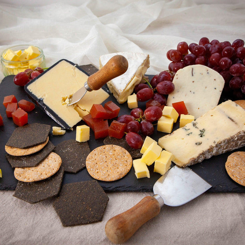 A cheese and charcuterie board with an assortment of cheeses, fruits, vegetables, and crackers, served with cheese knives.