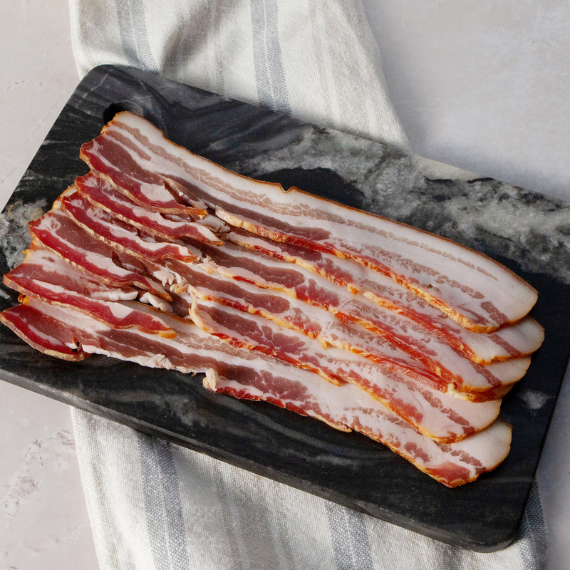 A pack of raw bacon strips laid out on a dark stone surface with a white and grey striped napkin beside it.